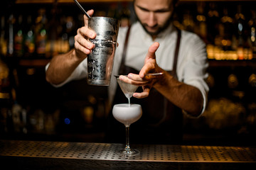 Professional male bartender pouring a white cocktail from the steel shaker to the glass through the sieve