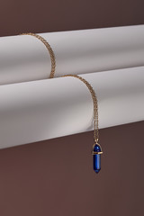 Close-up shot of a golden chainlet with a pendant in the form of screw with a fitted-in indigo-blue gemstone similar to a lazulite. The glazy gemstone is elongated and conical.  © RedUmbrella&Donkey