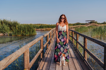 young beautiful woman standing on a wood pier in a natural park at sunset. Cute jack russell small dog looking at her owner.Tourism, love and travel concept