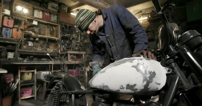 Focused adult guy in shabby clothes polishing plastered part of custom motorbike while working in workshop