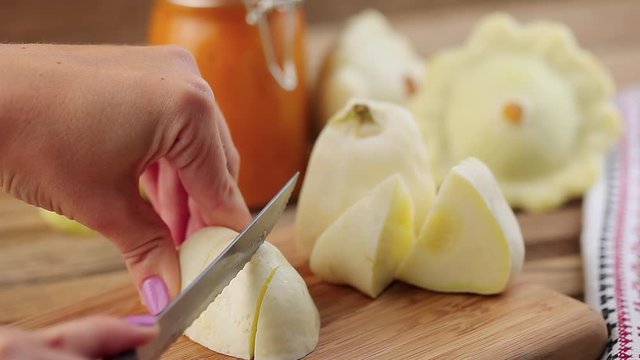 Female hands cutting a small white squash by knife on a wooden cutting board with a squash and glass jar on the background