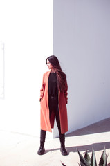Young long-haired woman is wearing a trench coat standing in front of a corner of a wall where one...