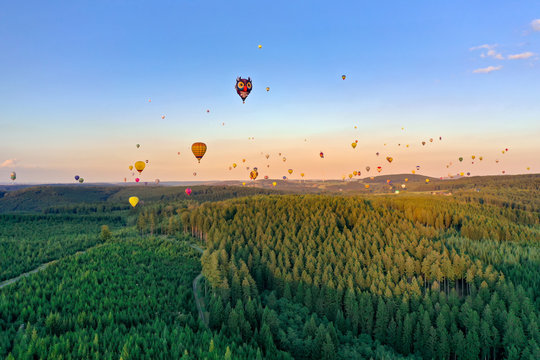 Colorful hot air balloons in different shapes fly over the forest. It is a coniferous forest in the Sauerland. The sun is almost setting, the sky is blue. Aerial shot.