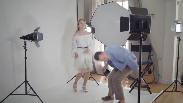 Professional male photographer dancing with a model in white dress before taking photos in the studio on white background. Photography, profession, photo session.