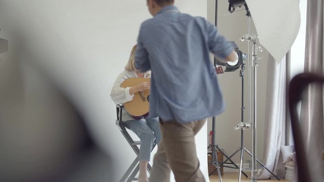 Backstage of the photo shoot. Famous professional photographer taking photos of young woman playing guitar while sitting on the chair on white background in the studio. Fashion studio photoshoot.