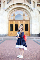 Back to school. The kid is getting ready for school. Primary school student. Girl with a backpack near the building outdoors. education. Little girl with a backpack holds stationery near the building