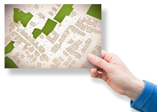 Imaginary cadastral map of territory with buildings, roads, land parcel and free green land available for building construction. Concept image with a female hand holding an postcard