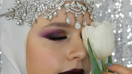 Professional model in a white image and a silver tiara with a bright multi-colored make-up called a smoky eye, posing in front of the camera in a model agency on a silver background. High fashion