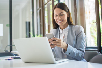 Beautiful young business woman using phone near laptop in office