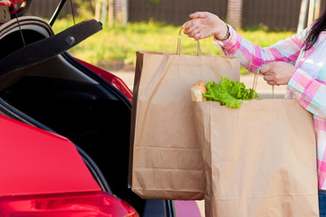 Young woman putting groceries from a supermarket in paper bags into the trunk of a car. Shopping...