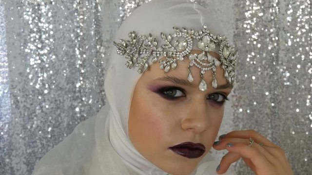 Face of model in a white image and a silver tiara with a bright multi-colored make-up called a smoky eye, posing in front of the camera in model agency on a silver background. High fashion