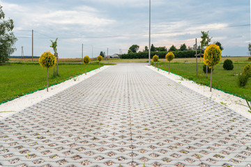 Entry for cars into the property made of openwork concrete pavement filled with colorful pebbles, around is green grass and small trees. - obrazy, fototapety, plakaty
