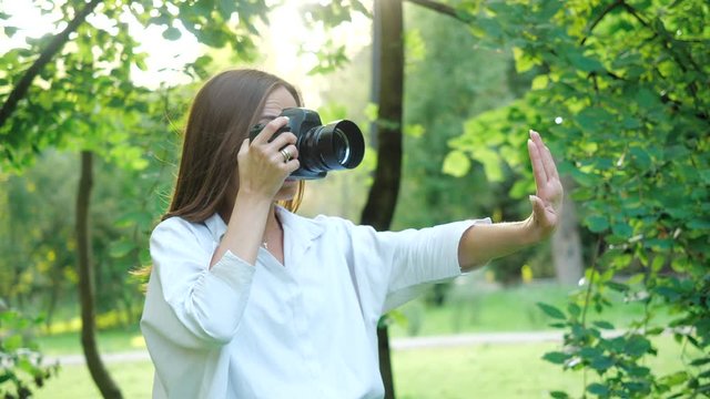 Pretty smiling and laughing girl photographer wearing white shirt is making photos in a park on a soft background of green foliage and spraying water.