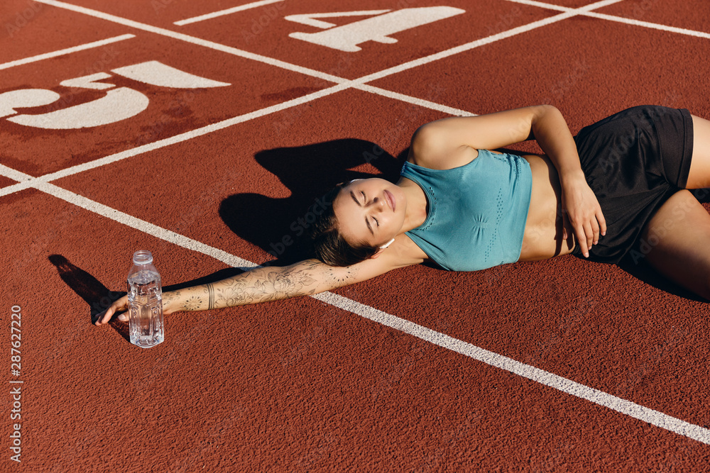 Wall mural beautiful athlete girl in sportswear dreamily lying on runner track with bottle after workout on cit