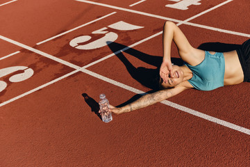 Attractive athlete girl in sportswear lying on runner track dreamily covering eyes with hand from sun after workout on city stadium