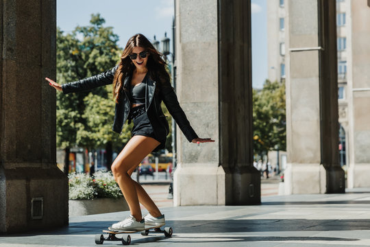 Beautiful young skater woman riding on her longboard in the city. Stylish girl in street clothes rides on a longboard. Skateboard, street photo, life style, freedom, happy face concept.