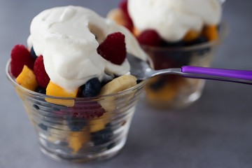 Fruit salad of banana, mango, blueberry and raspberry with whipped cream in glass glasses on a gray background.
