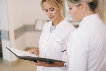Fototapeta premium Two women doctors standing with clipboard at hospital and discussing cases in modern white office. Girl doctor intern listens to the head doctor's instructions.