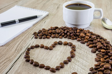 Heart made using coffee beans, cup of coffee on a wooden background, notebook and pen