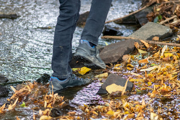 Junge mit schwarzer Hose und Wanderschuhen überquert einen Fluss über einen kleinen Damm aus Steinen und balanciert über glitschige Steine und Blätter und bekommt nasse Füße aber hat draußen viel Spaß