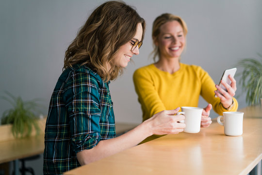 Two girlfriends sitting in coffee shop, drinking coffee, using smartphone