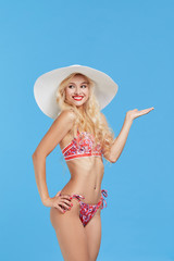 Portrait of a smiling and shocked attractive woman in bikini and hat points a hand to the right and up and looking at camera isolated over blue background. Travel and vacation concept. 