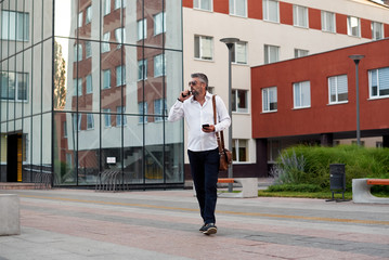 Middle-aged male businessman talking loudly on mobile phone and smoking a vape, blowing smoke. City and office building background