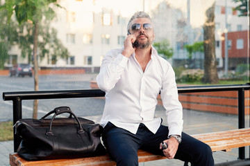 Middle-aged male businessman talking loudly on mobile phone and smoking a vape, blowing smoke. City and office building background