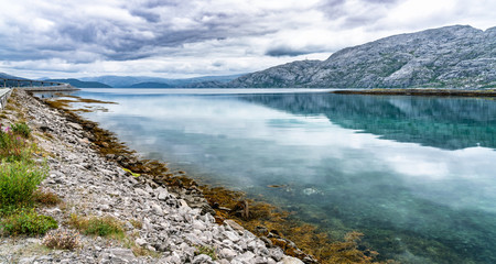 The clouds above fjord and mountains. Tides out but green and brown seagrass remains on the coast of Alsta Island. Close to Sandnessoen town, Norway