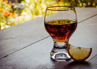 Brandy or cognac, rum, scotch in the glass with lemon on table. Wooden background texture. Brown strong alcohol drink for relaxation. Orange flowers.