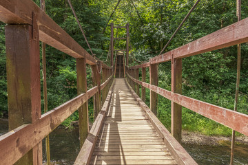Fototapeta na wymiar A small wooden suspension bridge passing through the river in the forest, which is mounted on cables against the background of deciduous trees.