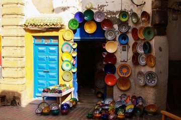 View on entrance of arabian shop with handmade colorful painted clay plates hanging on wall in bright sun and shade , Medina of  Essaouira, Morocco