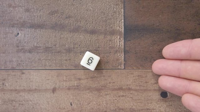 Anonymous hands rolls the number 6 on a six sided white die, on wood background.