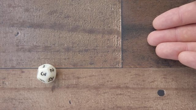 12 sided dice, hand rolls a 3 on brown floor or table, 4k flat lay, slow.