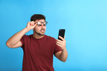 Young man with vision problems using smartphone on blue background, space for text