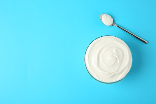 Glass bowl of sour cream and spoon on light blue background, flat lay. Space for text