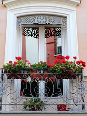 Beautiful balcony with white window frames and red flowers in the planters hanging on cast iron decorative railings 