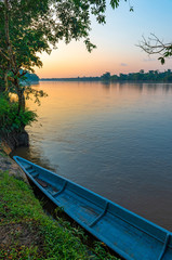 Vertical photograph of the Amazon Rainforest at sunset with a blue canoe in the foreground along...