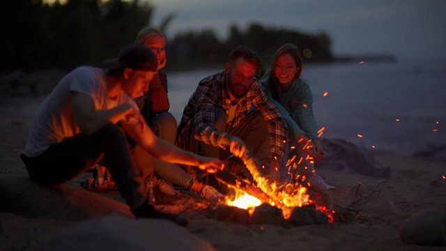 Four young adventure seekers, two men and two women, sitting together by campfire at night. Male friends adjusting woods to keep fire burning