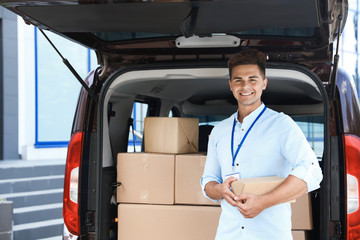 Young courier with parcel near delivery car outdoors