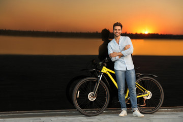 Fototapeta premium Handsome young man with bicycle near glass building at sunset. Space for text