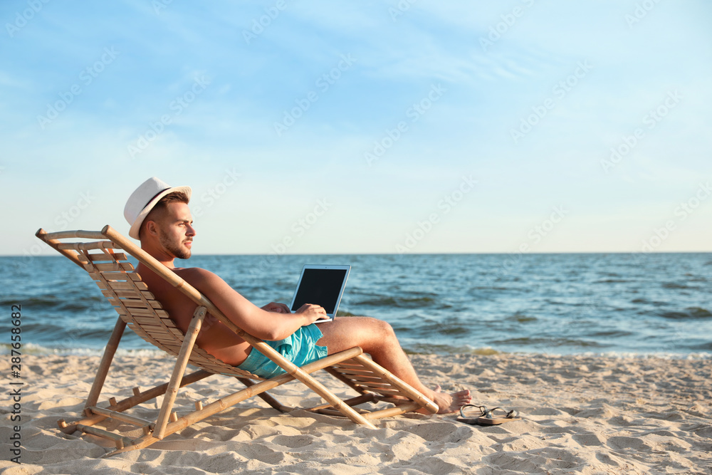 Wall mural Young man with laptop in deck chair on beach