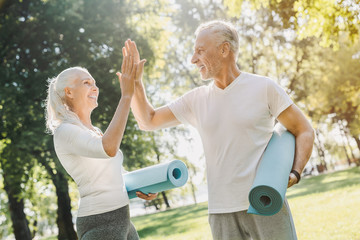 Mature couple standing with yoga carpet in hands in morning park before workout with high five each other
