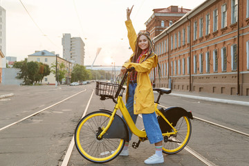 Young woman wearing yellow coat and colored pigtails, riding bike in city.