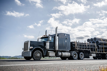 Black big rig semi truck transporting plastic pipes on step down semi trailer running on the road...