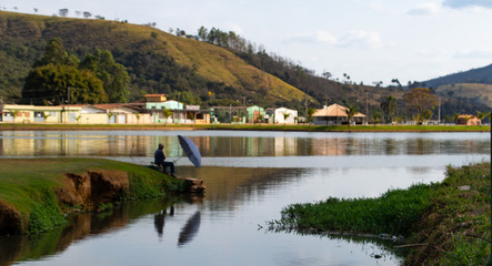 Fototapeta na wymiar a lone fisherman shading himself from the sun with an umbrella while fishing on Capitol Lake, Minas Gerais