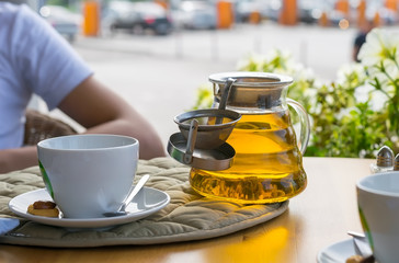 Brewed tea in a teapot on a table in an outdoors cafe on the background of tea cups, mobile phone, wallet and a man sitting at a table
