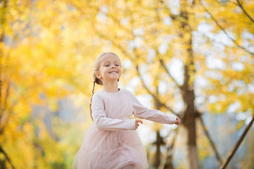 Happy stylish little girl with long blonde hair running in autumnal park with ginkgo trees. Autumn season, childhood magic, freedom, health concept