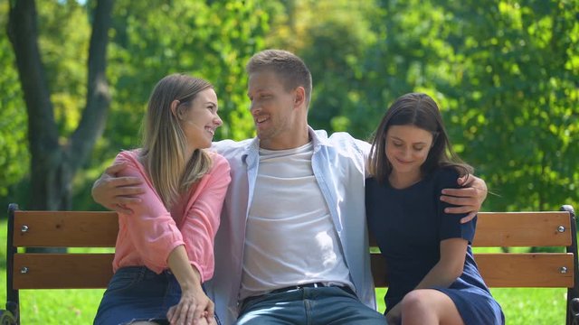 Handsome man resting on bench in park hugging two attractive girls, ladies man