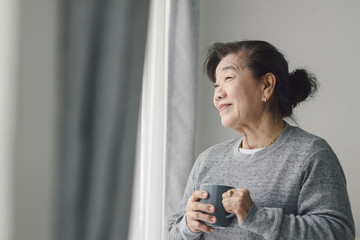 Asian senior woman drinking hot tea near window outdoor, lonely concept.